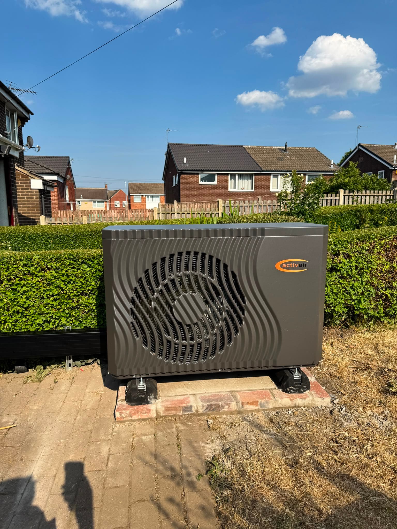 ActivAir monobloc air source heat pump installed on a brick plinth beside a neat garden hedge