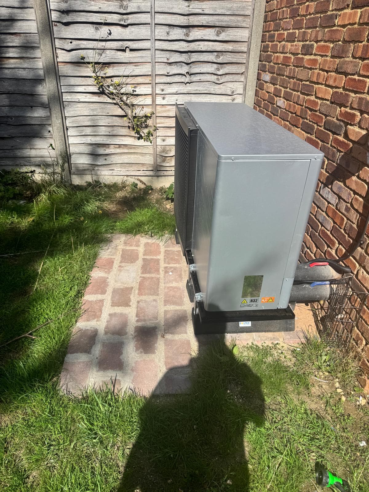 Grey R32 monobloc air source heat pump on a wall bracket above a block-paved base in a garden