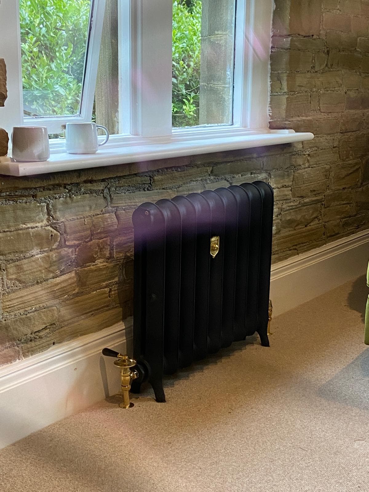 Black cast iron radiator with polished brass valves beneath a window on an exposed stone wall