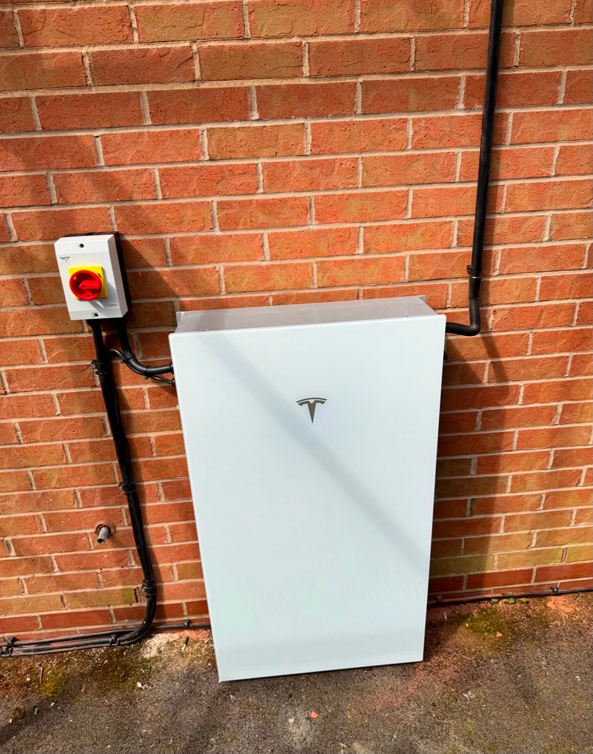 Tesla Powerwall home battery installed on a red brick wall with AC disconnect switch and black conduit